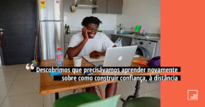 Imagem de jovem negro sentado a mesa de uma cozinha, expressa um sorriso ao falar pelo celular enquanto olha a tela de um notebook da Apple. Abaixo do centro da imagem a frase: “Descobrimos que precisávamos aprender novamente, sobre como construir confiança, a distância. No canto direito uma faixa na vertical da cor salmão, no canto inferior direito o logo do Voicers.